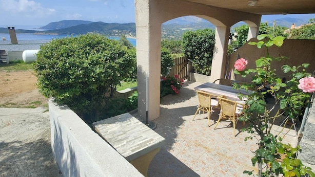 Un patio con mesa y sillas, vista al mar y jardín con arbustos y flores.
