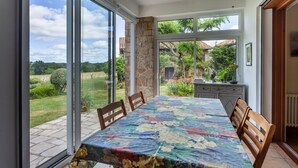 A dining area with a table covered in a floral tablecloth, surrounded by wooden chairs, and large windows offering a view of a garden and a house. - Guest Room 'Le Cèdre Bleu' with Shared Terrace & Garden, Wi-Fi (Rochechouart)