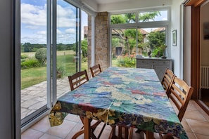 A dining area with a table covered in a floral tablecloth, surrounded by wooden chairs, and large windows offering a view of a garden and a house.