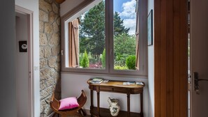 A room with a stone wall, a wooden chair with a pink cushion, a wooden table with books and a vase, and a view of a garden through a window. - Guest Room 'Le Cèdre Bleu' with Shared Terrace & Garden, Wi-Fi (Rochechouart)