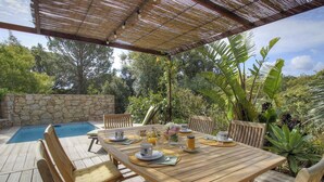 Un área de comedor al aire libre con piscina, muebles de madera y abundante vegetación.