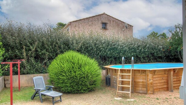 Una piscina de madera con una escalera, un tendedero metálico rojo, una silla de plástico gris y un arbusto grande.