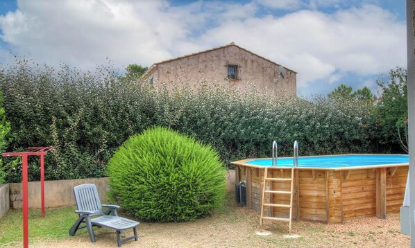 Une piscine en bois avec une échelle, un étendoir à linge en métal rouge, une chaise en plastique grise et un grand buisson.