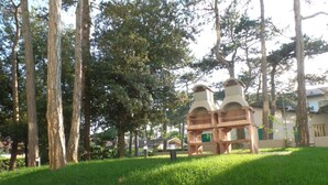 Un espacio cubierto de césped con dos estructuras de madera que parecen chimeneas, rodeado de árboles y un edificio al fondo.