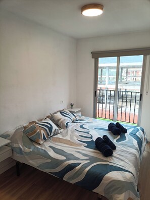 Ein Schlafzimmer mit Bett, Kissen, einem Nachttisch, einer kleinen Pflanze und Blick durch eine Schiebetür auf einen Außenbereich.