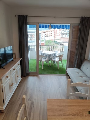 A living room with a TV, a sofa, a dining table, and a balcony with a view of buildings. - Apartment at the foot of Sierra de Irta, Hiking, Nature, Mountain and Sea. (Peñíscola)