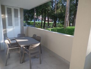 A balcony with a table and chairs, a white door with shutters, and a view of a green landscape with trees. - New flat for five guests in a residence with pool (Bibione)