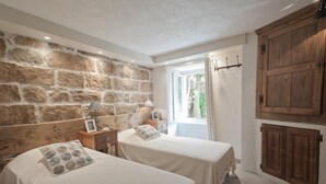 Een kamer met twee bedden, een stenen muur, een houten kledingkast en een raam met gordijnen.
