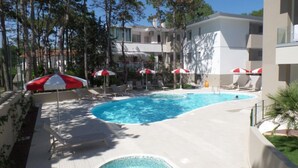 A swimming pool with lounge chairs and umbrellas, surrounded by trees and a building with balconies. - New flat in a residence with pool and jacuzzi (Bibione)