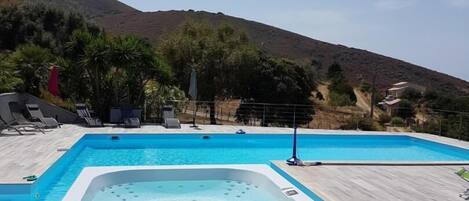 A swimming pool with a hot tub, lounge chairs, and a mountainous backdrop.
