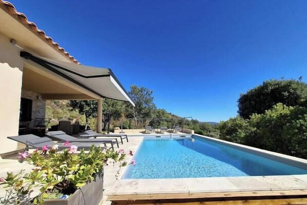 Ein Pool mit Liegestühlen und einer Pergola, umgeben von viel Grün und blauem Himmel.