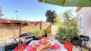 Een terras buiten met een houten terras, rieten meubels, een tafel met een rode placemat, een barbecue en potplanten.