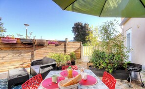 An outdoor patio with a wooden deck, wicker furniture, a table with a red placemat, a grill, and potted plants. - Le 28081 - ALATA - Gîte (ALATA)