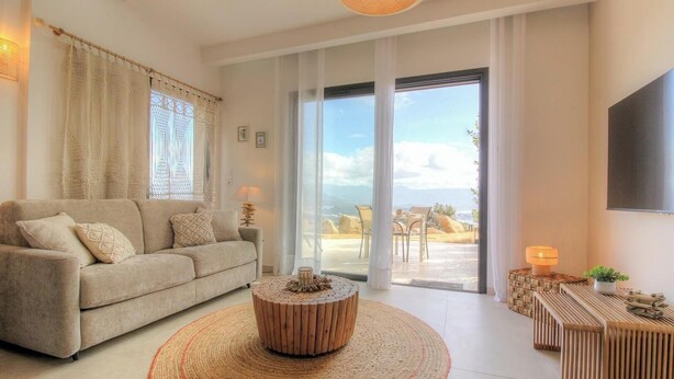 Ein Wohnzimmer mit einer Couch, einem runden Holztisch und Blick ins Freie durch große Glasüren.