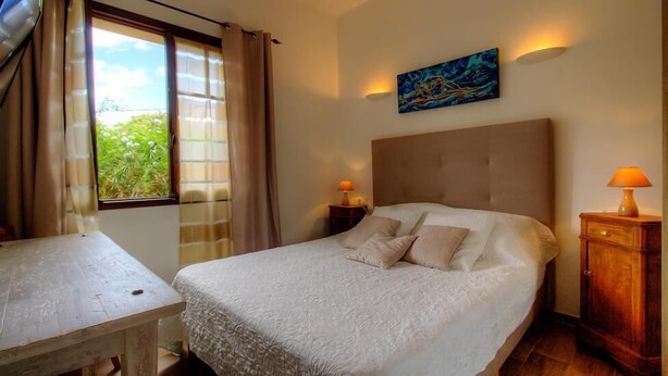 Un dormitorio con cama, una mesa, una mesita de noche, una lámpara y un cuadro en la pared.