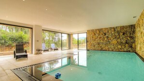 Una piscina interior moderna con paredes de piedra, ventanas amplias y una zona de estar con sillones.