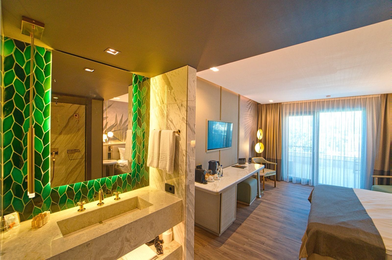 A hotel room with a large mirror, a vanity with a green mosaic backsplash, a bed, a desk with a flat-screen TV, and a large window with curtains.