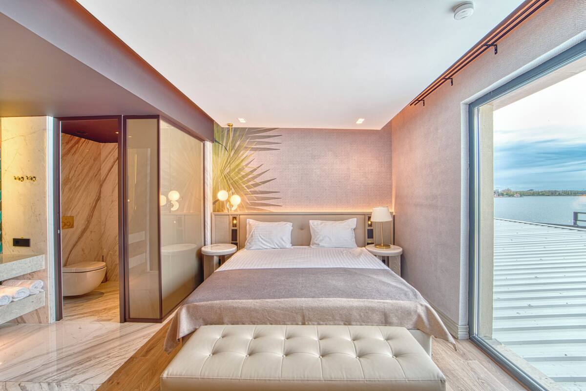 Une chambre moderne avec un grand lit, des tables de chevet, une cabine de douche en verre et une vue sur l’eau.