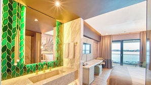 Una habitación de hotel moderna con una cama grande, un escritorio, un baño con salpicadero de mosaico verde y vistas al agua.
