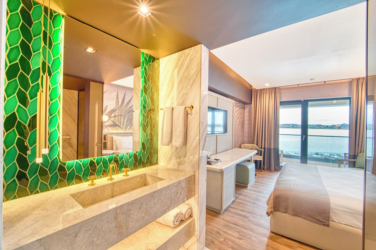 Une chambre d’hôtel moderne dotée d’un grand lit, d’un bureau, d’une salle de bain avec un mur recouvert de mosaïques vertes et offrant une vue sur l’eau.