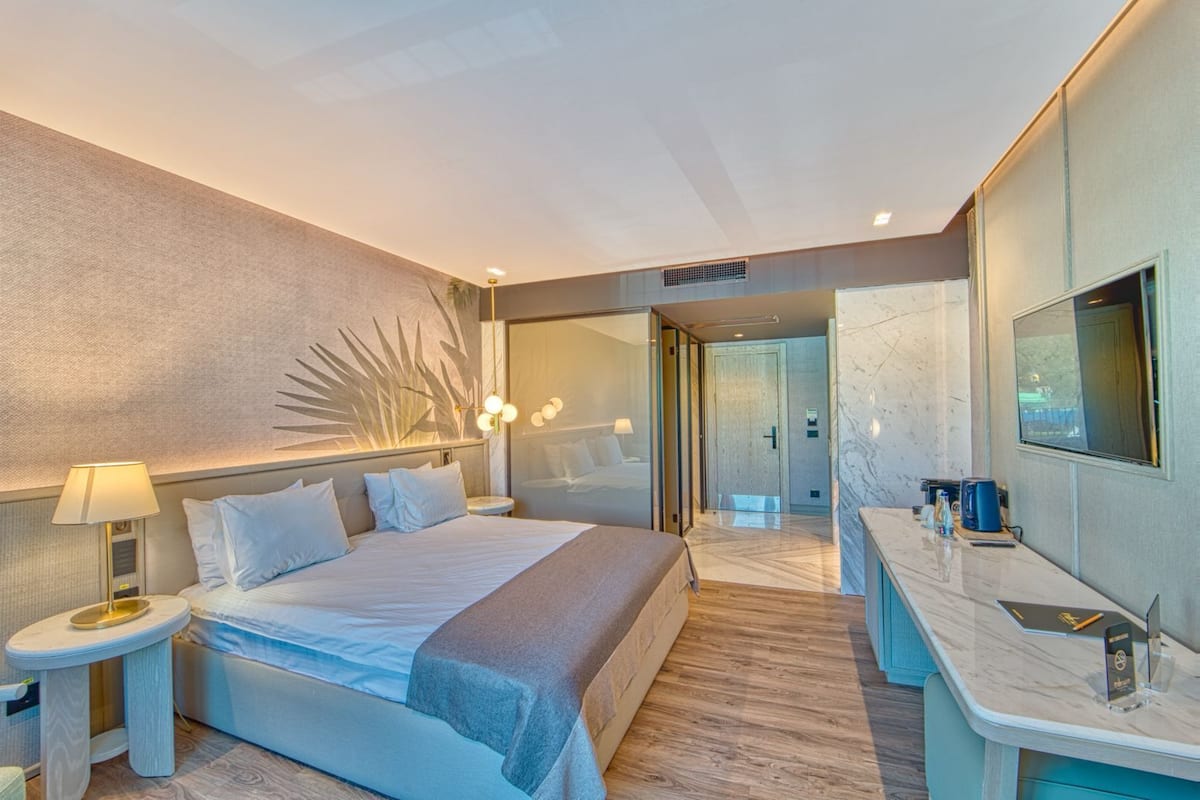 Une chambre d’hôtel moderne, dotée d’un grand lit, de tables de chevet, d’une télévision et d’une salle de bain visible à travers une porte en verre.