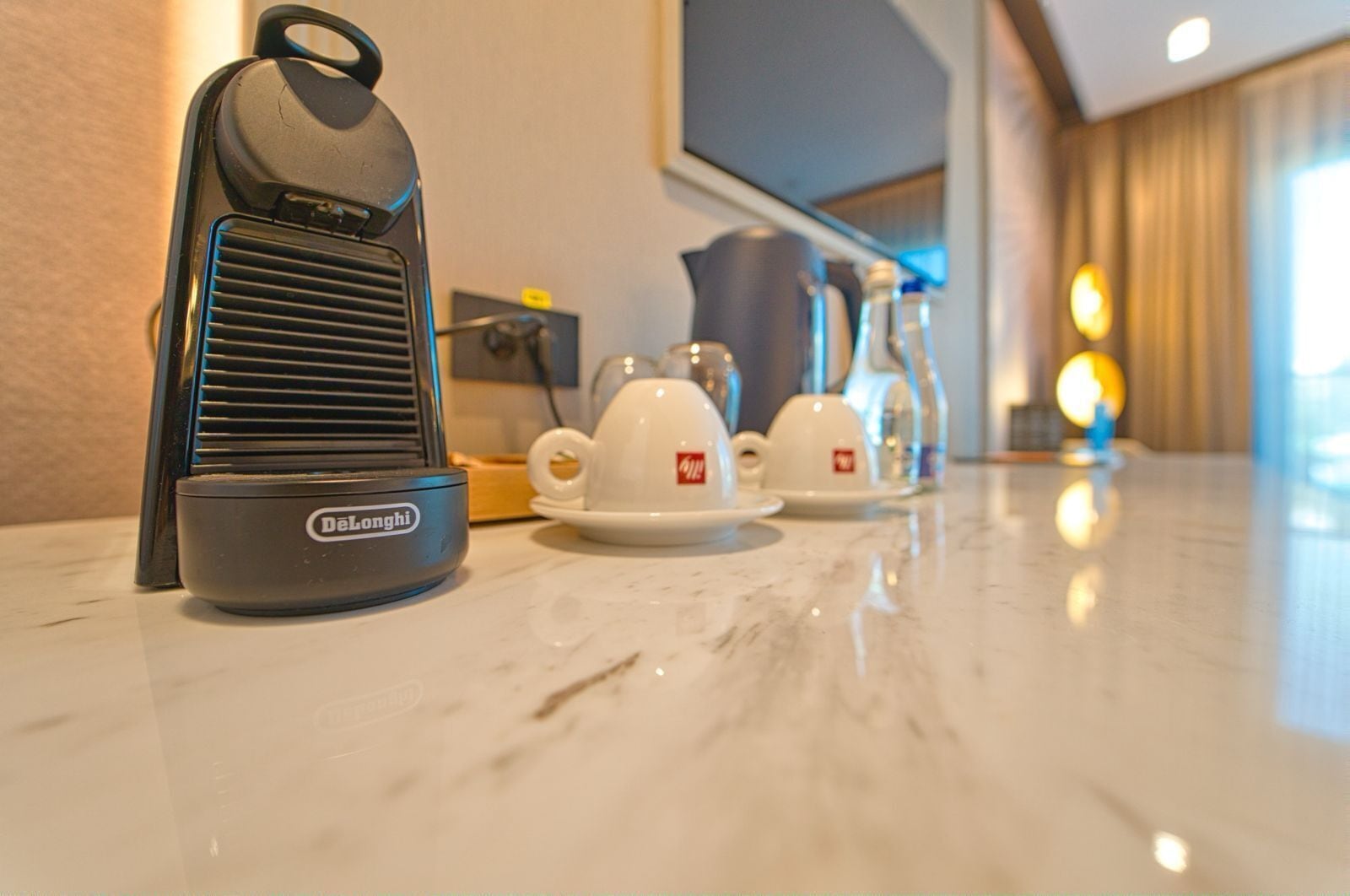 Un calentador portátil DeLonghi sobre una encimera de mármol, con dos tazas y platillos blancos al fondo.
