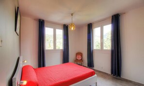 Ein Schlafzimmer mit einem Bett, zwei Fenstern, einem Sessel und einer Pendelleuchte.