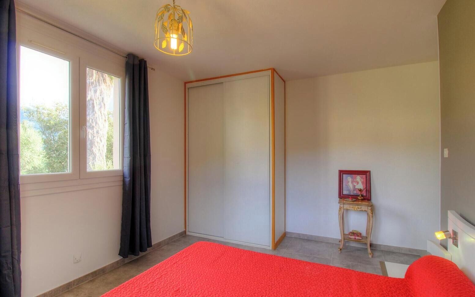 A bedroom with a bed, a window with curtains, a door, a small table with a picture, and a light fixture.