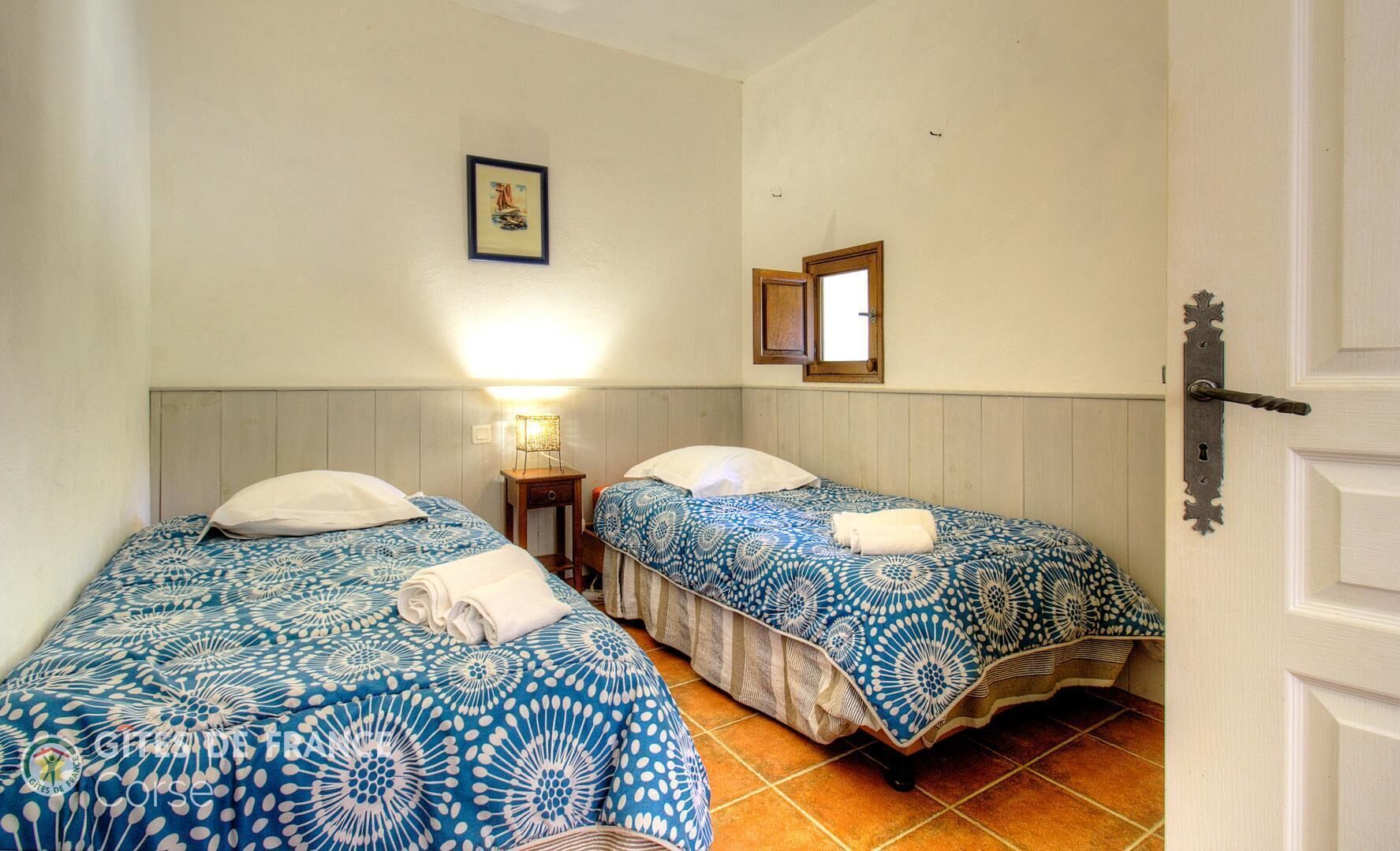 A room with two beds, a nightstand, a wall-mounted lamp, a framed picture, and a door with a decorative handle.