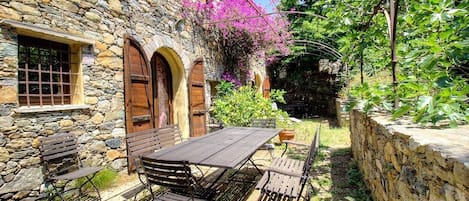 A stone building with a patio table and benches, surrounded by greenery and a stone wall.