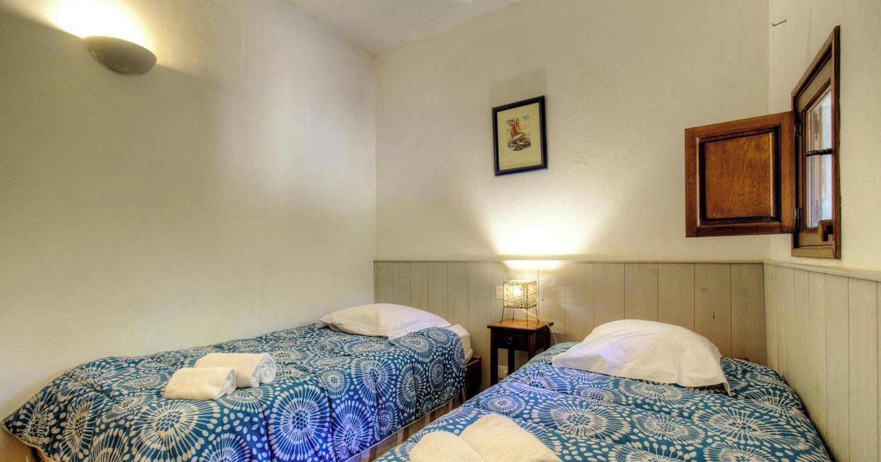 A room with two beds, a small table with a lamp, a framed picture on the wall, and a wooden cabinet.