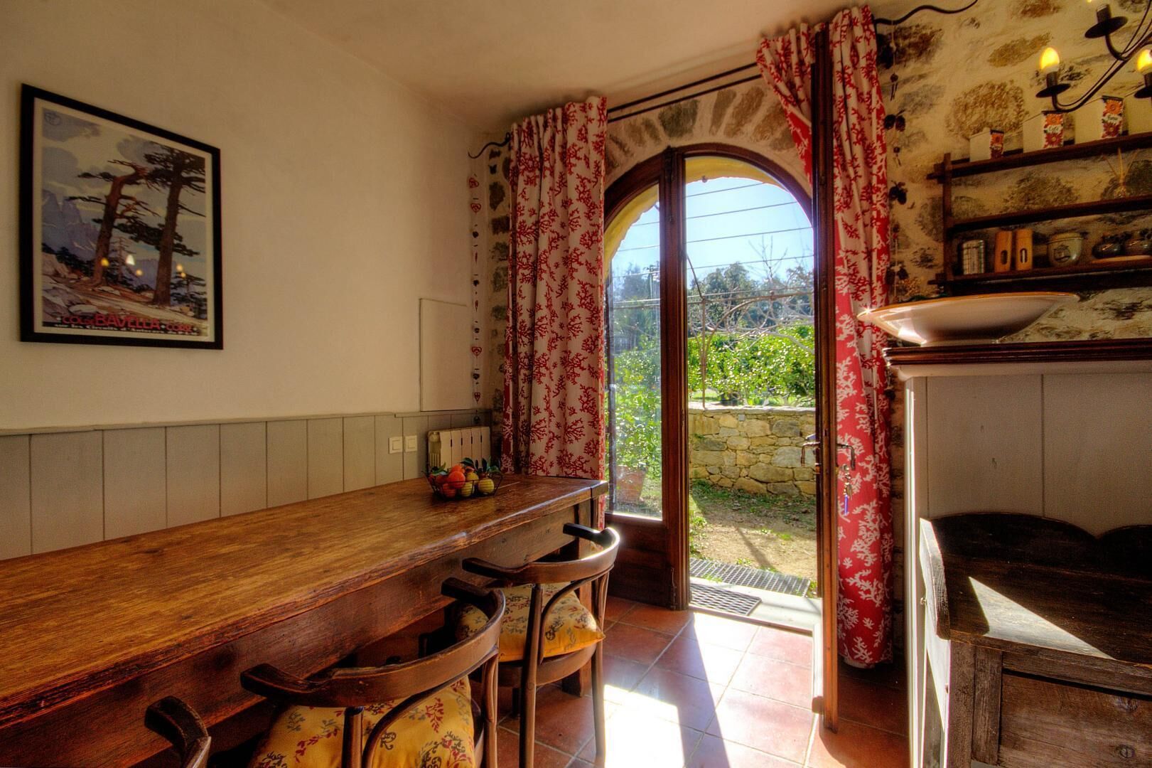 Eine Küche mit Holztisch, Stühlen, einem Esstisch, einem Fenster mit Vorhängen und einer Steinsäule im Freien.