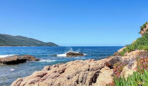 A coastal landscape with rocky terrain, clear blue water, and distant hills. - Villa Crique d'ANCONE (CALCATOGGIO)