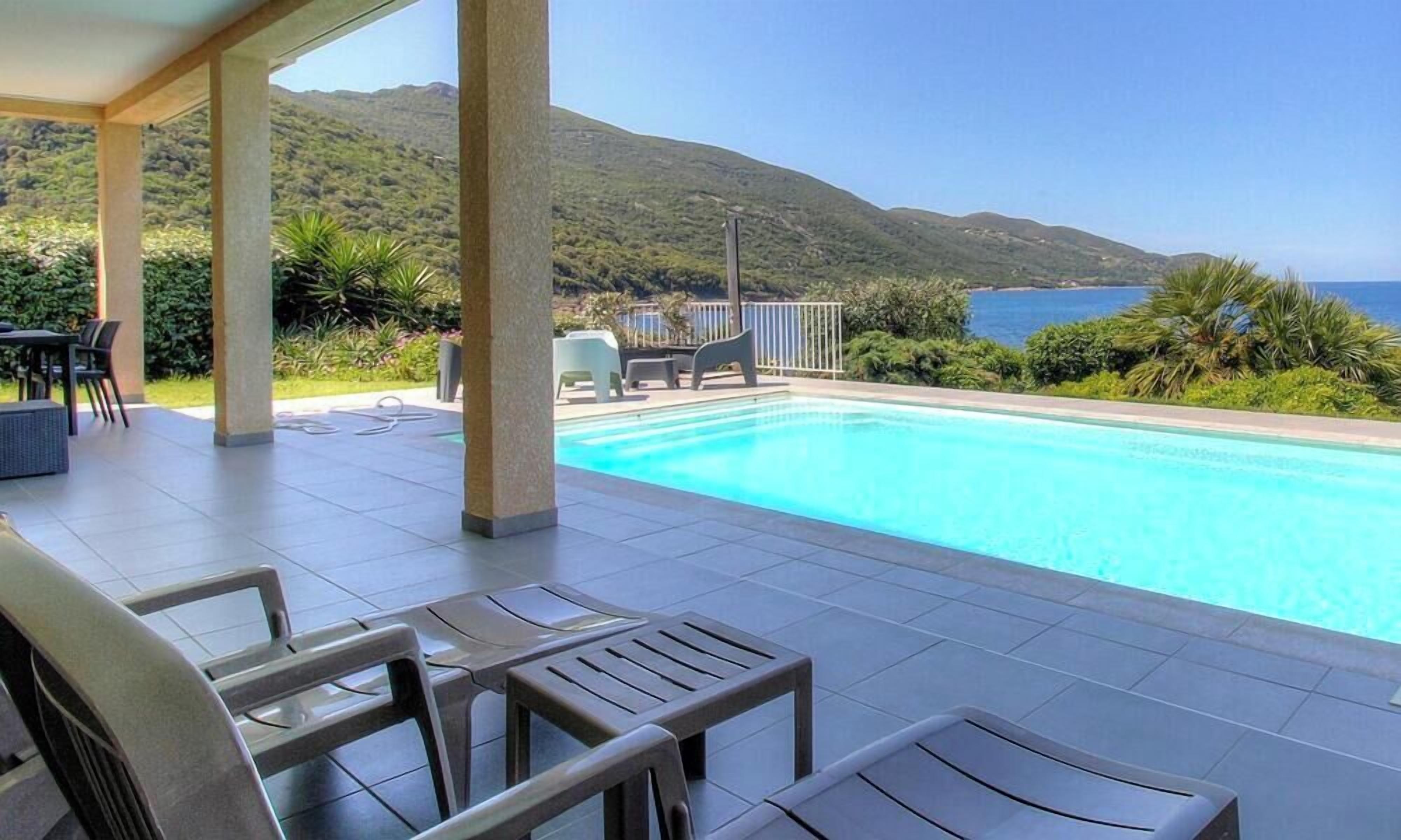 A pool with a view of the sea and mountains, outdoor furniture, and a covered patio area.