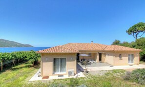 Una casa de dos pisos con techo de tejas, un patio con mesa y sillas, y vista al mar.