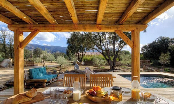 Eine Holz-Pergola mit Pool, Sitzgelegenheit und einem Tisch, gedeckt für das Frühstück.
