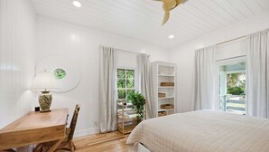 Ein Schlafzimmer mit einem Holztisch, einem Bett, einem Fenster mit Vorhängen und Blick auf Grün.