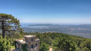 Un edificio de piedra con techo plano, rodeado de árboles verdes y frondosos, con vistas a un paisaje costero y colinas lejanas.
