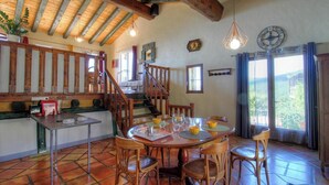 Una cocina rústica con vigas de madera, un comedor con una mesa redonda y sillas, y una escalera que lleva a un altillo.