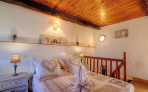 Un dormitorio con techo de madera, cama con almohadas estampadas con corazones, lámparas de noche y un pequeño estante con objetos decorativos.