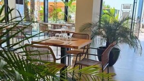 A spacious indoor area with a dining table and chairs, large windows, and potted plants. - Zena Hotel (Rrashbull)