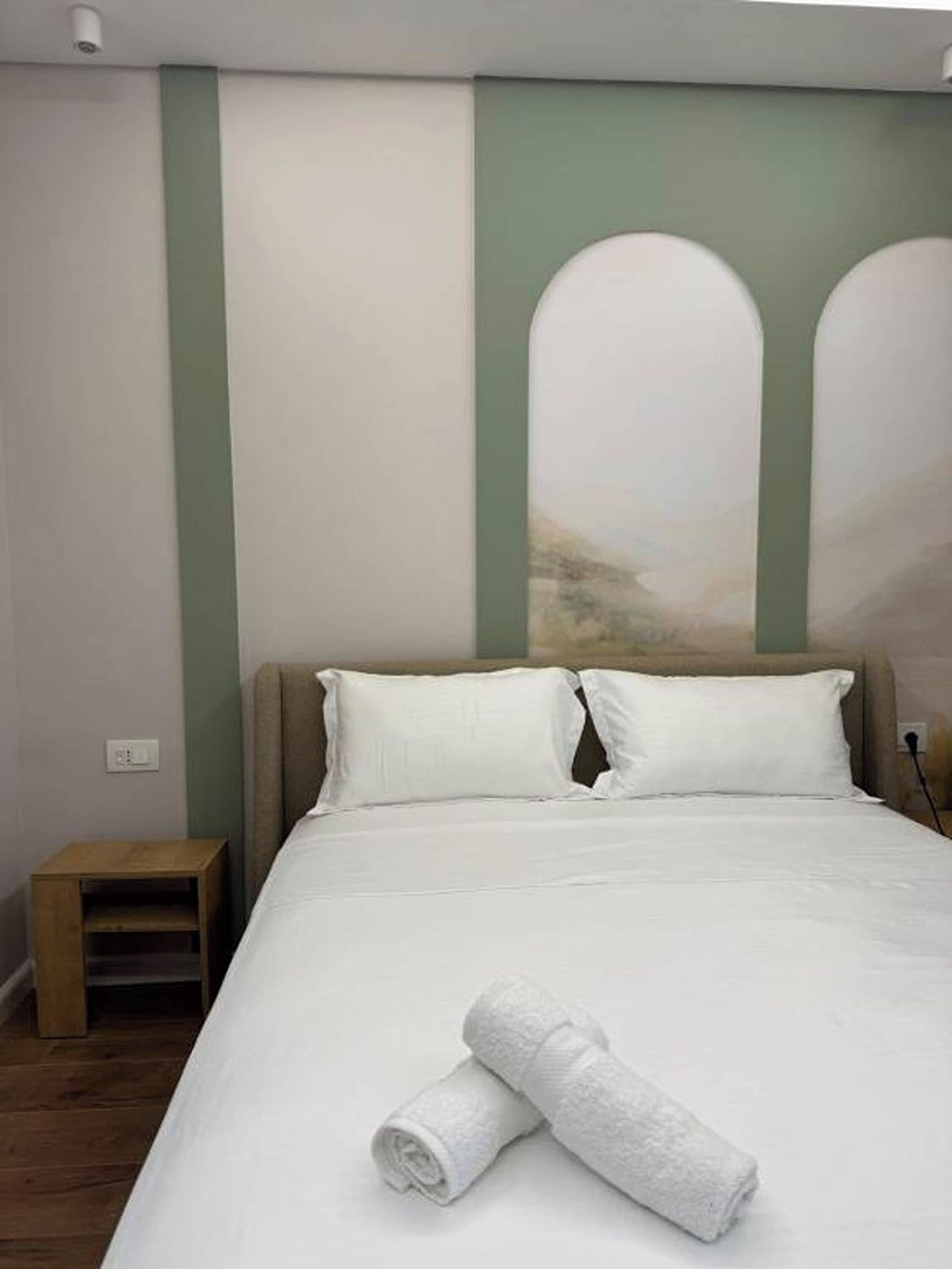 A hotel room with a bed, pillows, a nightstand, and a wall with a painting.