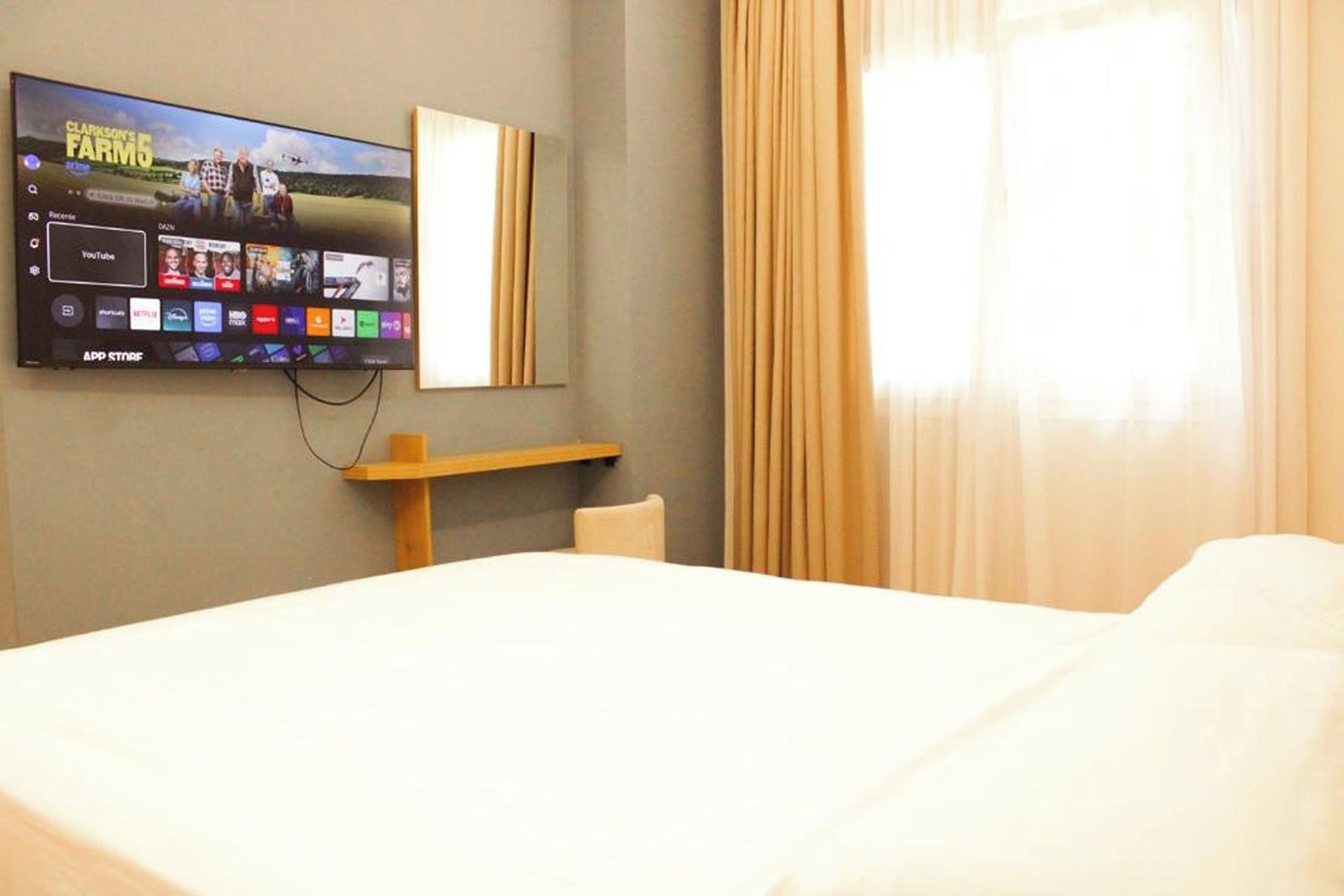 A hotel room with a bed, a TV mounted on the wall displaying various channels, a wooden shelf, and a window with curtains.