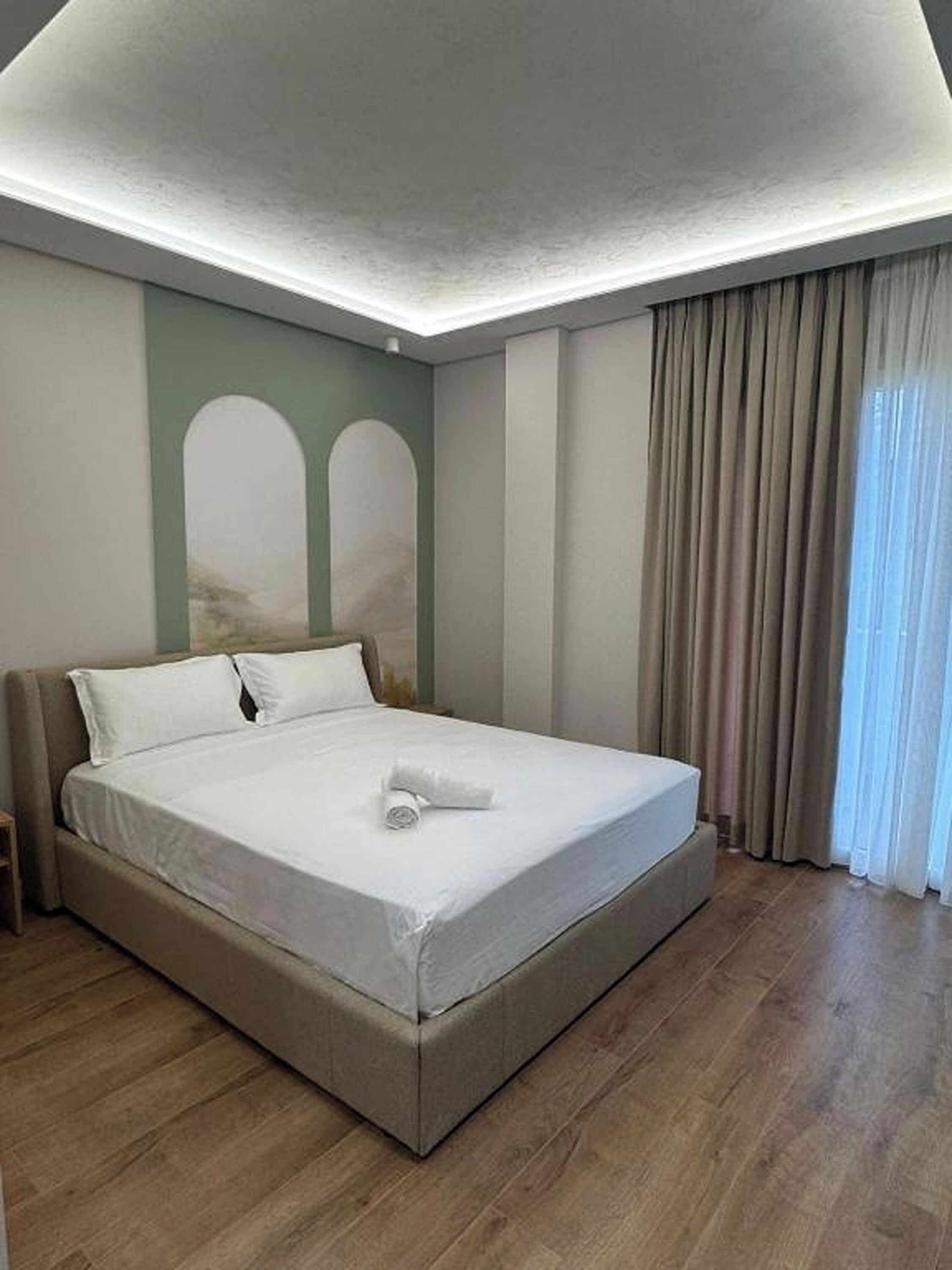 A modern hotel room with a large bed, a headboard, a nightstand, and a window with curtains.