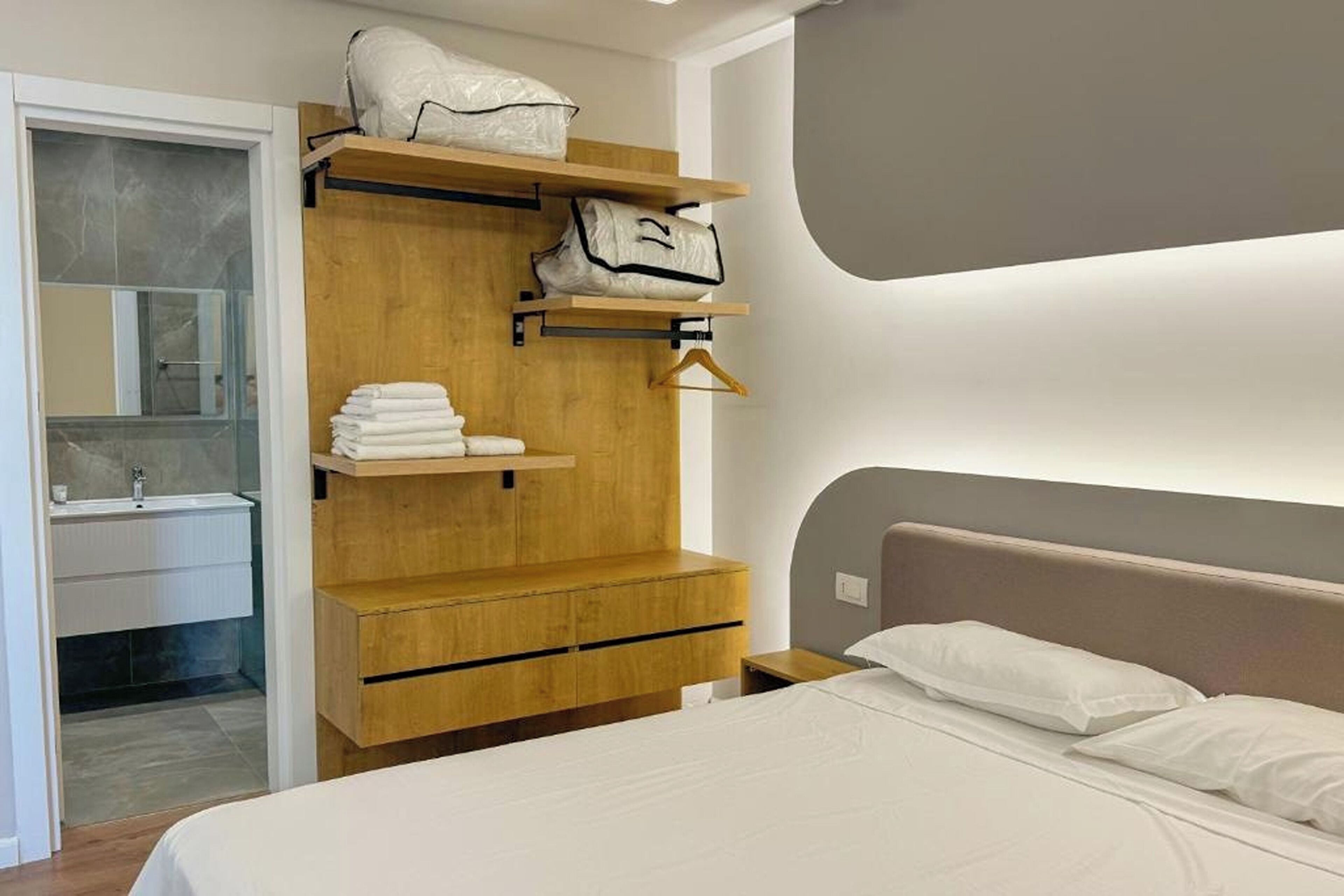 A modern bedroom with a wooden wardrobe, a bed with white linens, and a bathroom with a glass shower enclosure.
