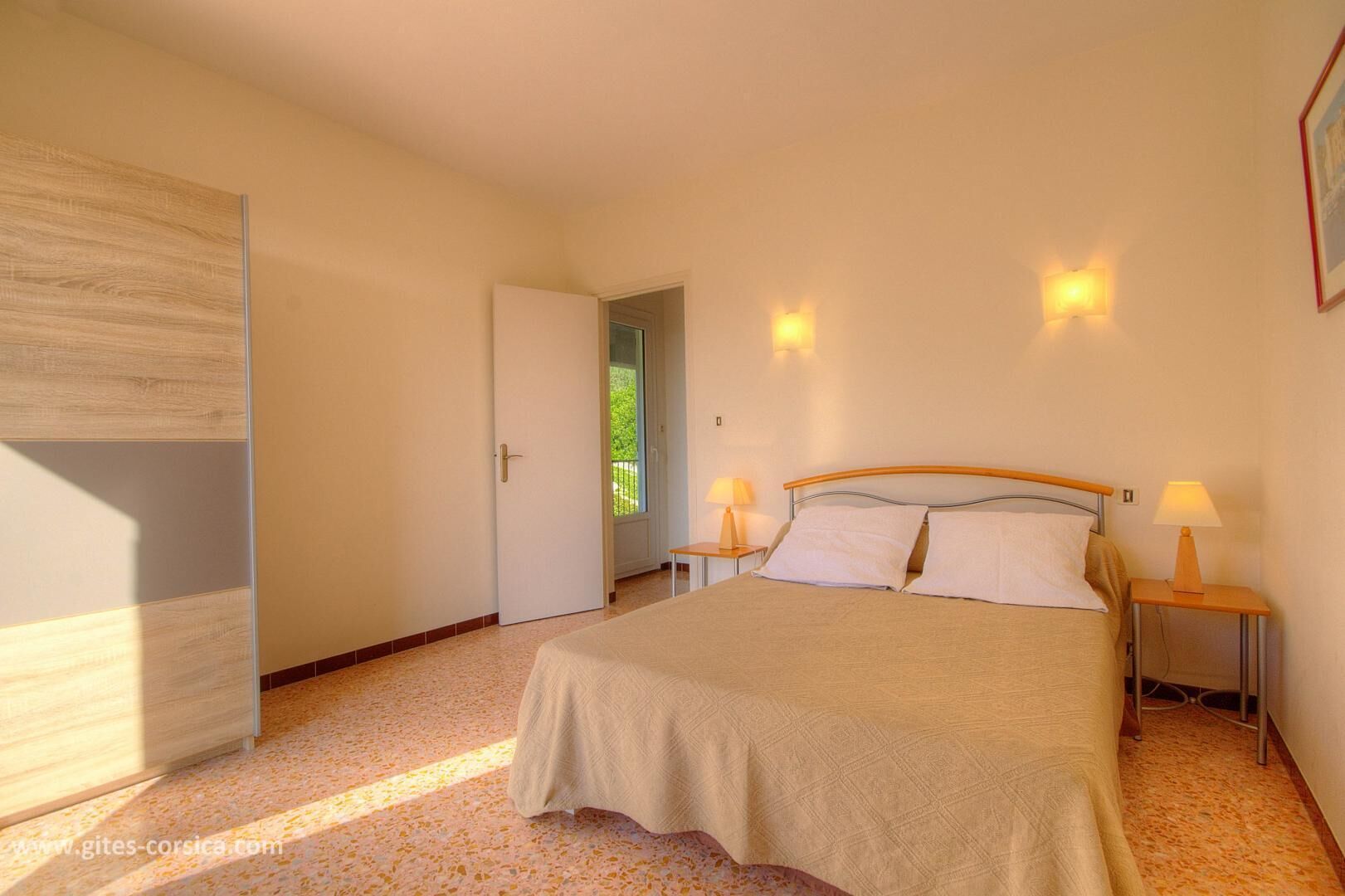 Un dormitorio con una cama, dos lámparas de noche, una puerta y vistas a la vegetación exterior.