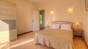 Ein Schlafzimmer mit einem Bett, zwei Nachttischlampen, einer Tür und Blick auf Grünfläche.