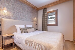 A bedroom with a large bed, a nightstand, a wall-mounted lamp, and a window with a view of a building.