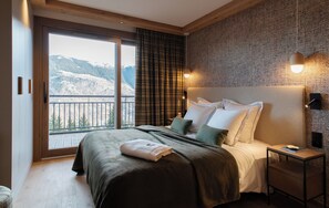 A bedroom with a large bed, a nightstand, a lamp, and a view of mountains through a balcony door.