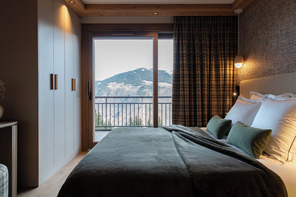 A bedroom with a bed, a view of mountains, a balcony, and a wooden ceiling.